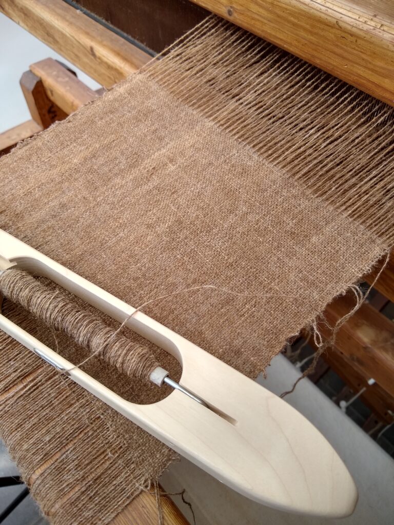 brown wool sample of tabby weaving on the loom with a boat shuttle in pale wood on top.